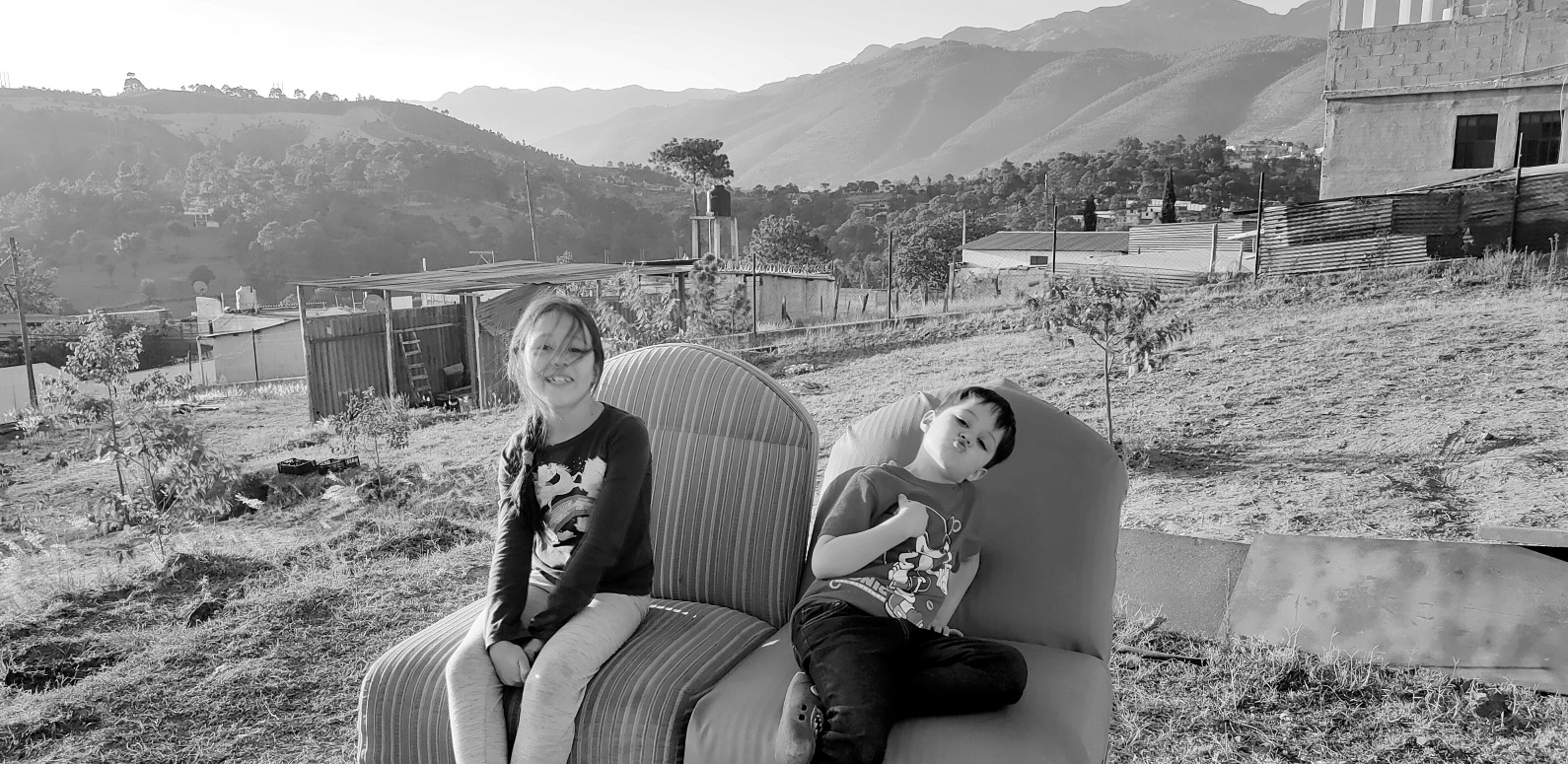 Two children sitting on an old armchair on a cleared building plot, with the Cuchumatanes mountains behind them, near the Guatemala-Mexico highlands