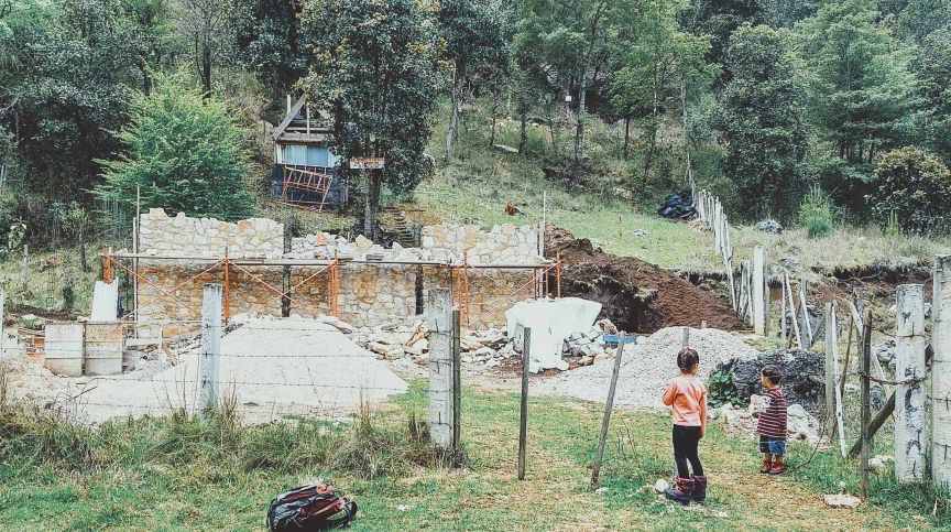 A half-built stone-walled house with concrete columns, scaffolding, gravel piles and materials on a wooded hillside near San Cristóbal de las Casas, Chiapas.