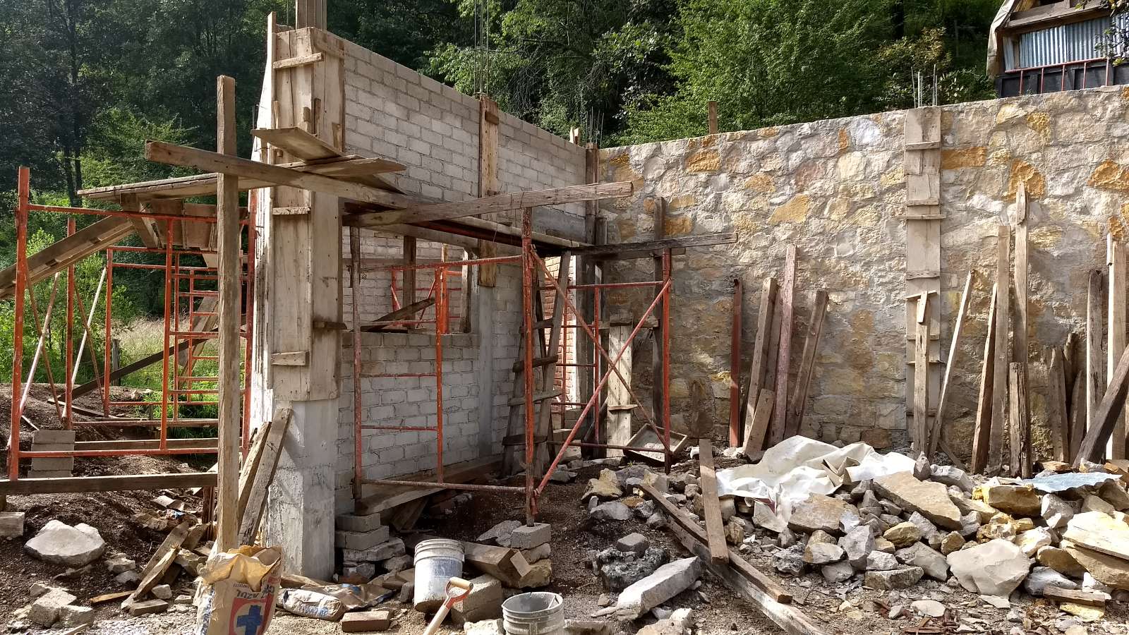 Tabique walls of a downstairs kitchen under construction with wooden scaffolding, in front of a stone containing wall on a self-built house site in Chiapas