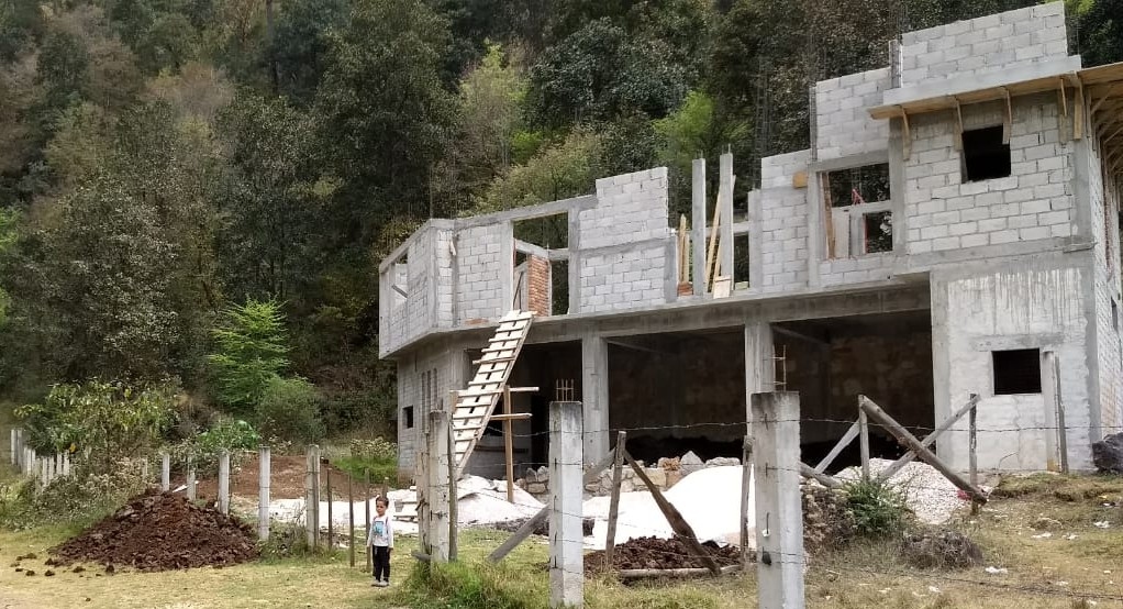 Two-storey concrete-block house shell under construction on a forested plot in San Cristóbal de las Casas, with a wooden site staircase up to the first floor and building materials in the yard