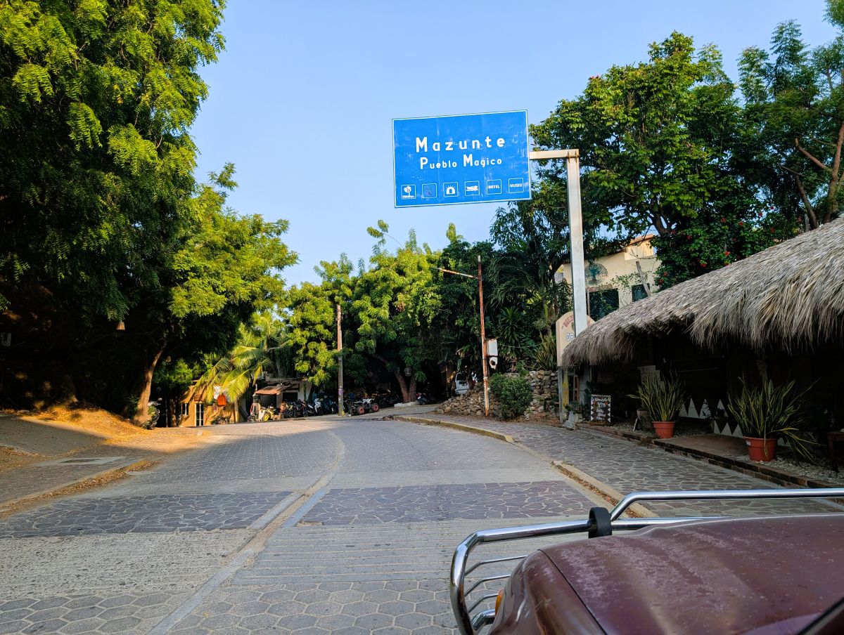 Blue Mazunte Pueblo Mágico road sign above the street into town, trees on the left and a palapa-roofed building on the right, seen over the bonnet of the car