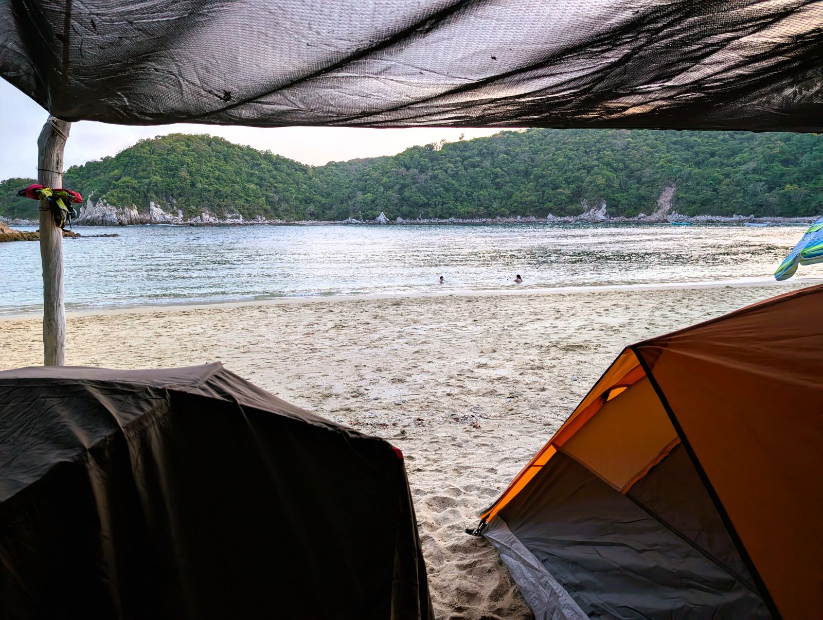 View out from under a tarp shelter at Playa Riscalillo, Oaxaca — two tents on the sand, calm bay water and a forested headland beyond